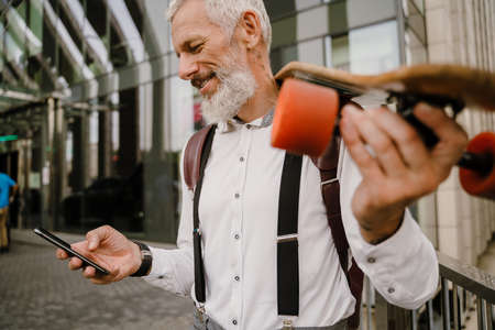 Grey mature man using cellphone while standing with skateboard at city streetの写真素材