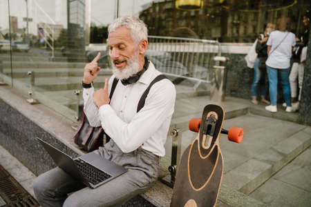 Grey mature man gesturing while working with laptop while sitting at city streetの写真素材