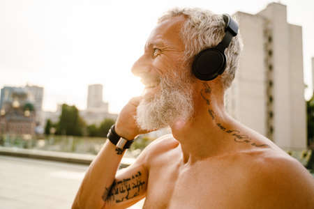 Shirtless mature man listening music with headphones while standing outdoorsの写真素材