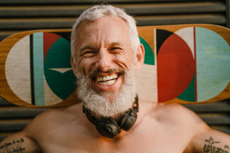 Shirtless mature man laughing while standing with skateboard outdoorsの写真素材
