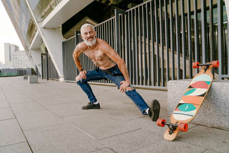 Adult white shirtless man doing workout by his skateboard outdoors