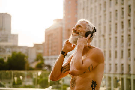 Shirtless mature man listening music with headphones while standing outdoorsの写真素材
