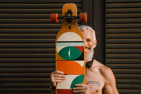Shirtless mature man smiling while standing with skateboard outdoors