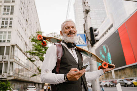Grey mature man using mobile phone while standing with skateboard at city streetの写真素材
