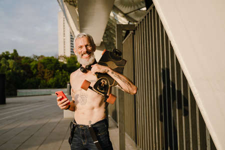 Adult white shirtless man holding cellphone while standing with skateboard on his shoulders outdoors