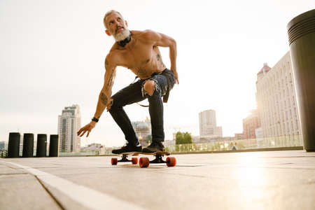 Shirtless mature man with beard skateboarding on parking outdoorsの写真素材