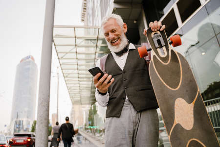 Grey mature man using mobile phone while walking with skateboard at city streetの写真素材