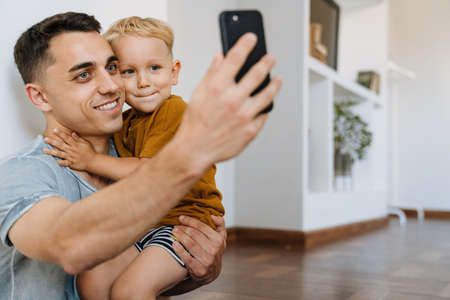 Young father and son smiling and taking selfie photo on mobile phone at homeの写真素材