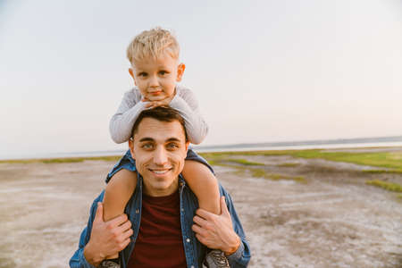 Young white happy father carrying little son on his shoulders while walking outdoors