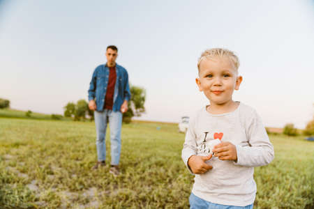 Blonde boy looking at camera while standing with her father on summer fieldの写真素材