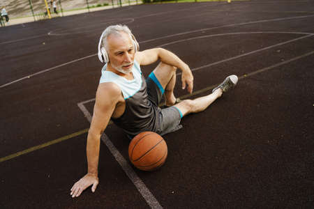 Grey senior man listening music while sitting with ball on playgroundの写真素材