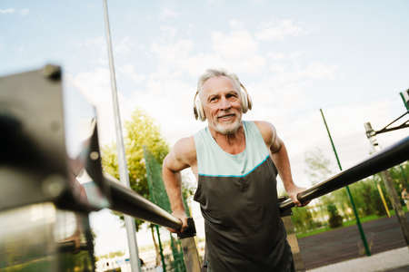 Grey senior sportsman listening music while working out on parallel bars at sports ground