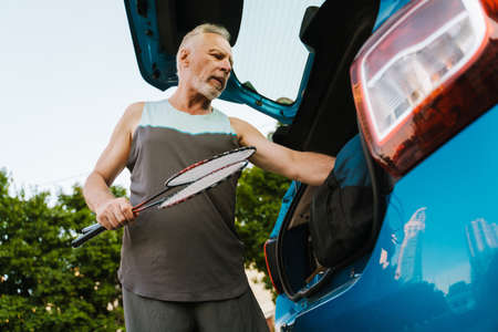 Grey senior sportsman getting badminton rackets out of bag in trunk outdoorsの写真素材