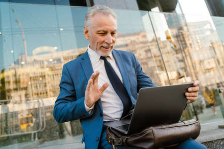Senior man dressed waving hand and using laptop while sitting by building at city street