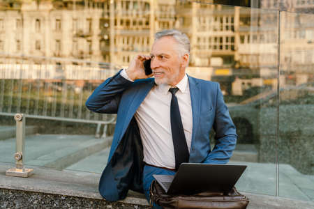 Senior man dressed in suit talking on cellphone while working with laptop on city streetの写真素材