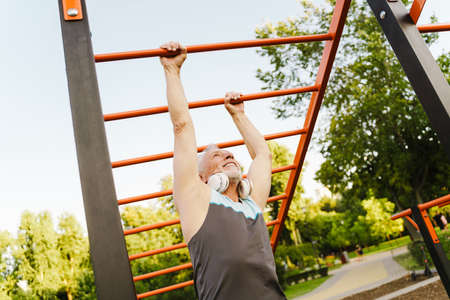 Grey senior man listening music while working out on equipment on sports ground