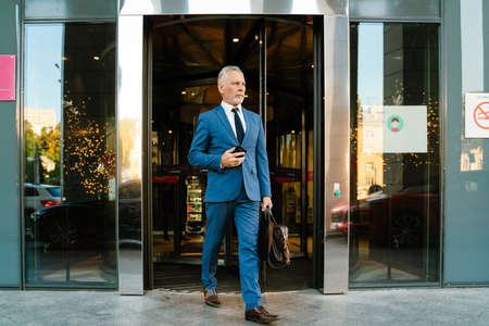 Senior man dressed in suit using mobile phone while coming out door of building