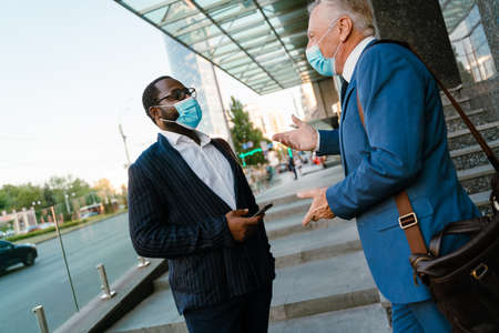 Multiracial men in face masks talking and gesturing while standing by building outdoorsの写真素材