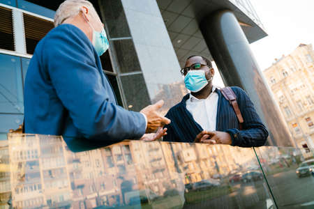 Multiracial men in face masks talking and gesturing while standing by building outdoorsの写真素材