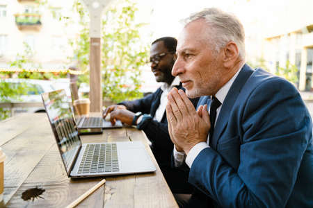 Multiracial men gesturing and working with laptops while sitting in cafe outdoors