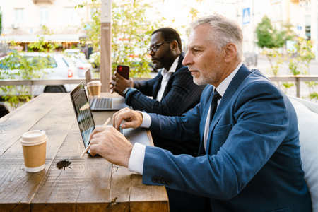 Multiracial men working with laptops while sitting in cafe outdoorsの写真素材