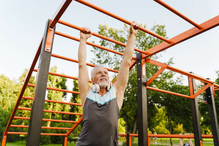 Grey senior man listening music while working out on equipment on sports ground