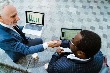 Multiracial men talking and handshaking while working with laptops outdoors
