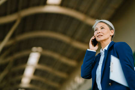 Grey asian woman talking on cellphone while walking with clipboard outdoorsの写真素材