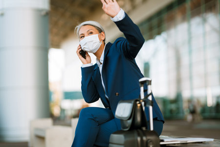 Grey woman in face mask waving hand while talking on cellphone by airport outdoorsの写真素材