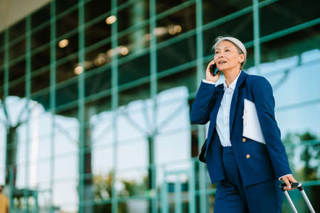 Grey asian woman talking on cellphone while walking with suitcase outdoorsの写真素材