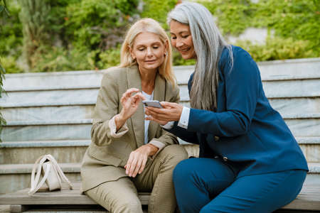 Mature multiracial businesswomen using cellphone while sitting on bench outdoorsの写真素材