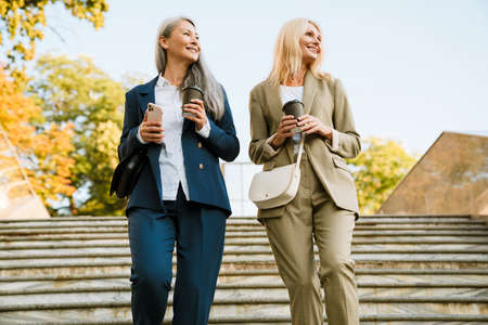 Mature multiracial businesswomen talking and drinking coffee while going down stairs outdoorsの写真素材