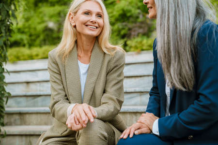 Mature multiracial businesswomen talking and smiling while sitting on bench outdoorsの写真素材
