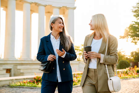Mature multiracial businesswomen talking and drinking coffee during walking outdoorsの写真素材