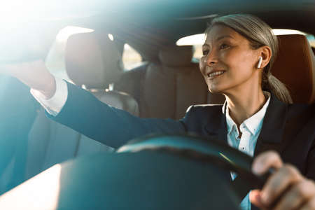 Mature asian woman using earphones and looking in rear-view mirror while driving car