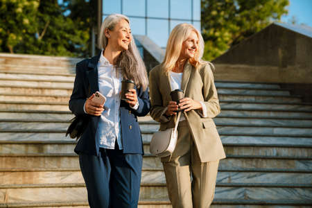 Mature multiracial businesswomen talking and drinking coffee while going down stairs outdoorsの写真素材
