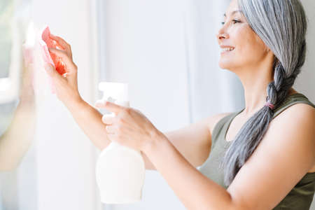 Mature asian woman with grey hair cleaning window with spray at homeの写真素材