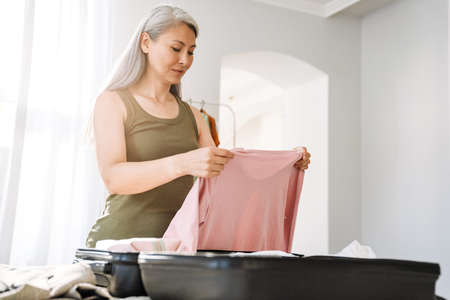 Mature asian woman with grey hair packing suitcase on bed at homeの写真素材
