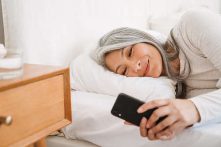 Grey asian woman using mobile phone while lying in bed after sleep at homeの写真素材