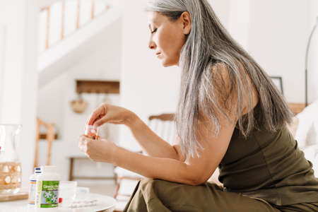 Mature asian woman taking her medication while sitting on couch at homeの写真素材