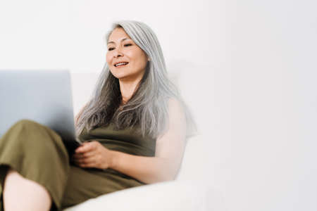 Mature asian woman working with laptop while sitting on couch at homeの写真素材