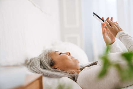 Grey asian woman using mobile phone while lying in bed after sleep at homeの写真素材