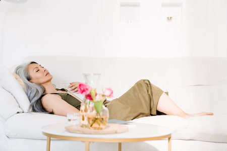 Grey asian woman sleeping with book while lying on couch at homeの写真素材