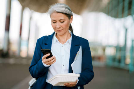 Grey asian woman holding food box and using mobile phone at airport parkingの写真素材