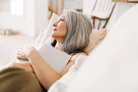 Grey asian woman looking aside while lying with book on couch at homeの写真素材