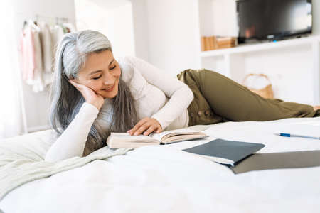Grey asian woman smiling and reading book while lying on bed at homeの写真素材