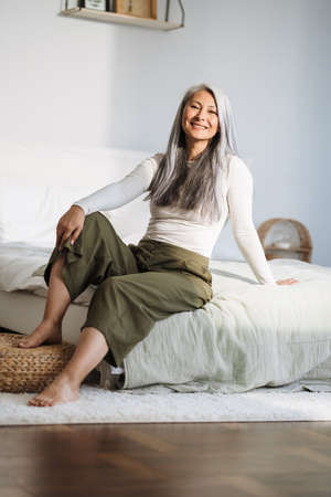 Mature asian woman with grey hair smiling while sitting on bed at homeの写真素材