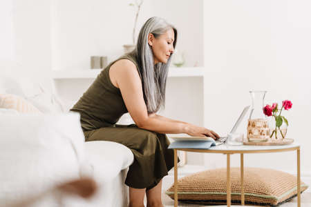 Mature asian woman working with laptop while sitting on couch at homeの写真素材