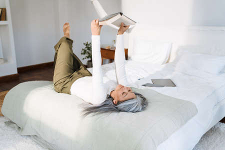 Grey asian woman reading book while lying on bed at homeの写真素材