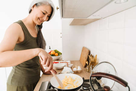 Mature asian woman smiling while cooking scramble eggs in kitchen at homeの写真素材